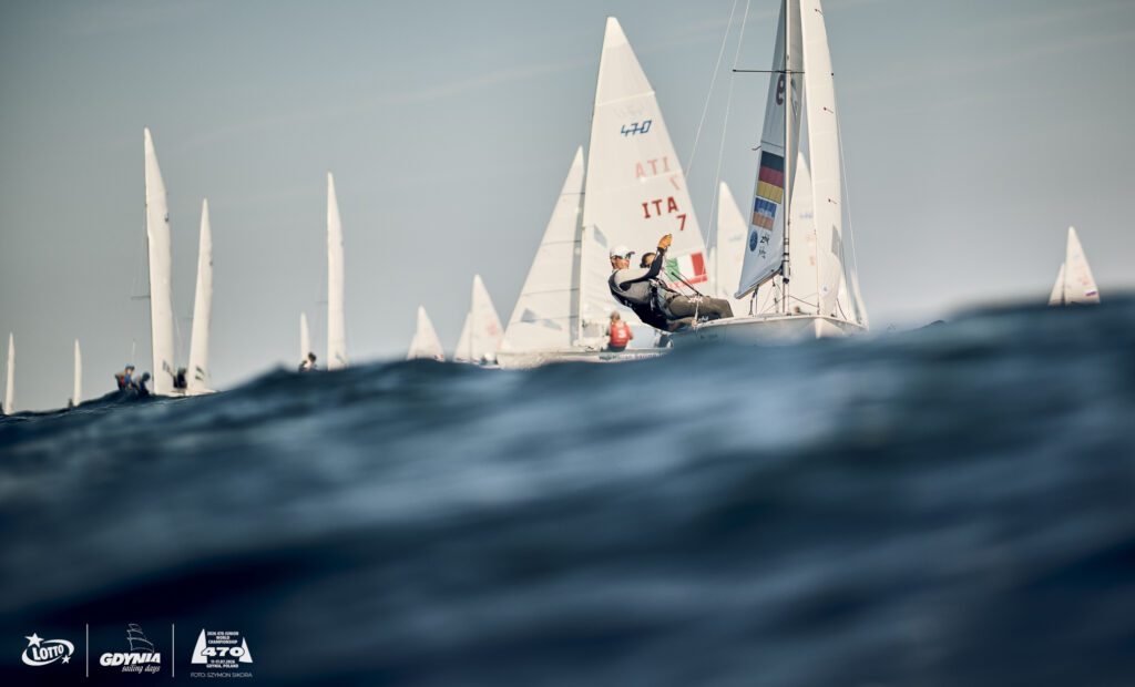 Eine große Welle und mehrere 470er Jollen, die eine Regatta segeln.