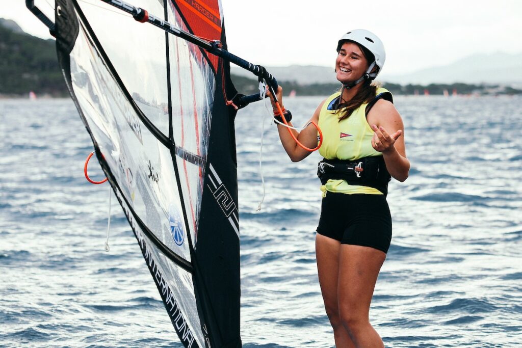 Eine Frau mit einem weißen Helm und einem gelben Trikot steht auf einem Windsurfboard.