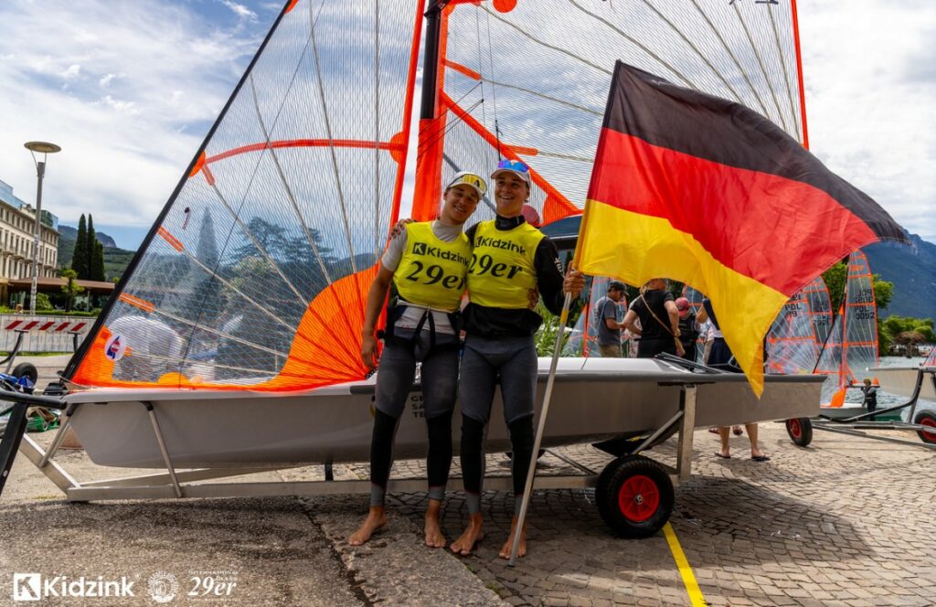 Zwei Segler mit einem gelben Trikot jubeln vor einem Boot mir durchsichtigen Foliensegeln: