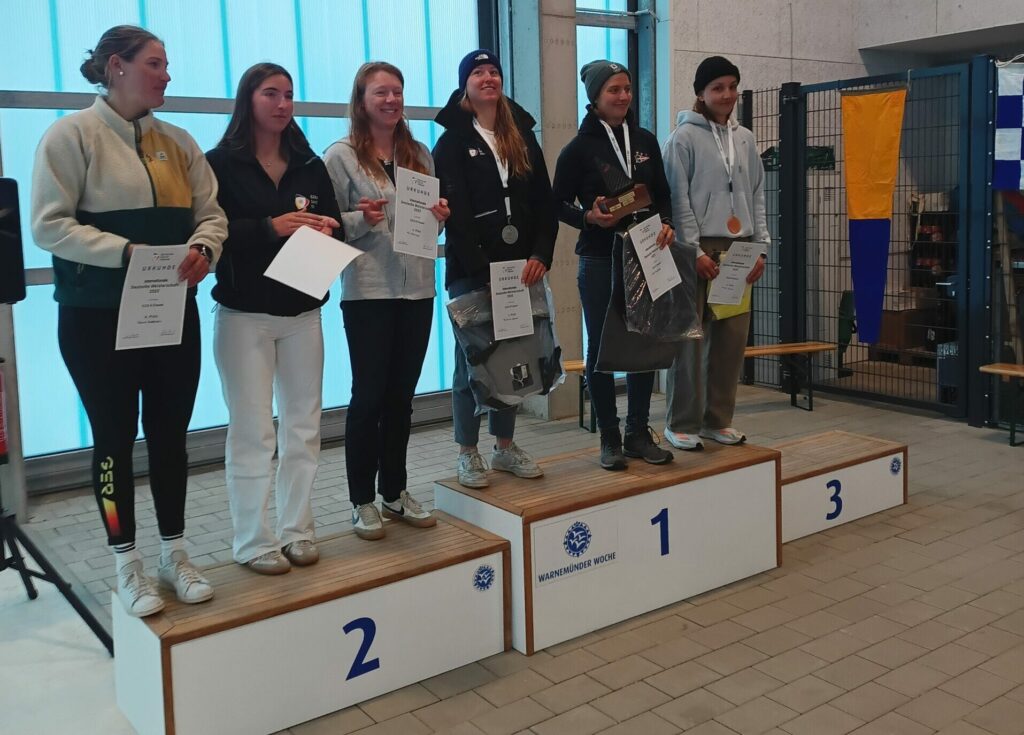 Sechs Frauen auf einem Siegerpodest, sie tragen Sportbekleidung und zum Teil Mützen.