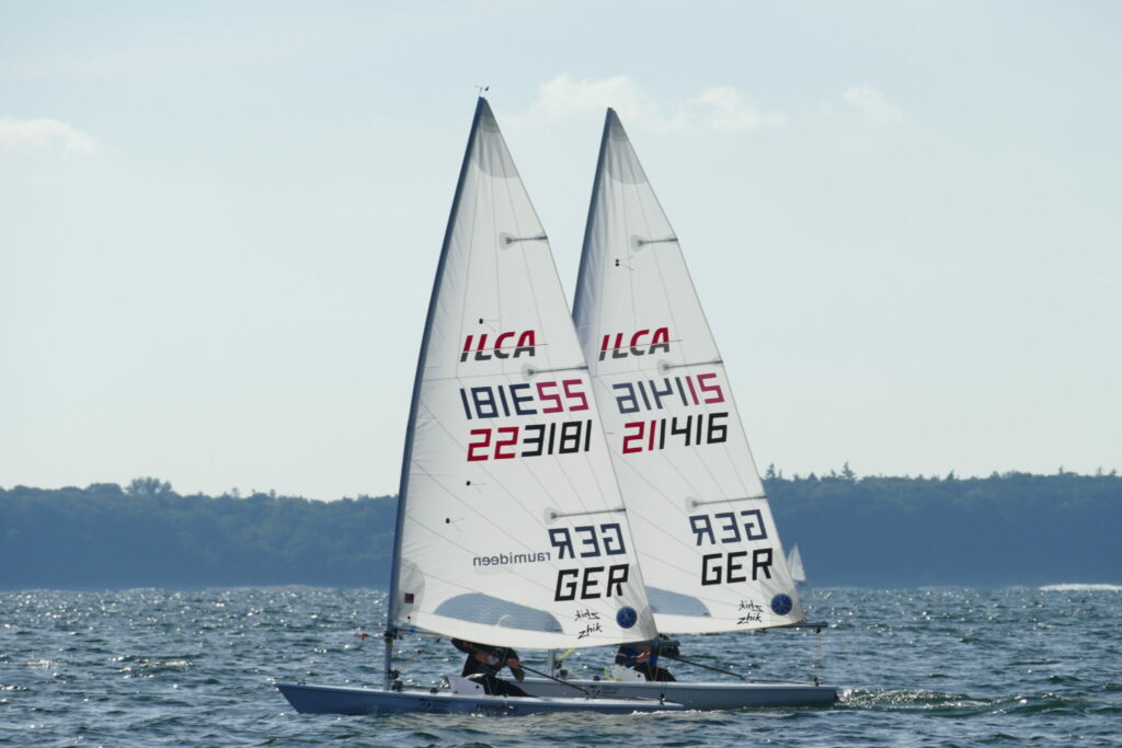 Zwei ILCA Jollen, auch als Laser bekannt, segeln nebeneinander am Wind, die Segler sind verdeckt, man kann die Segelnummern sehr gut erkennen.