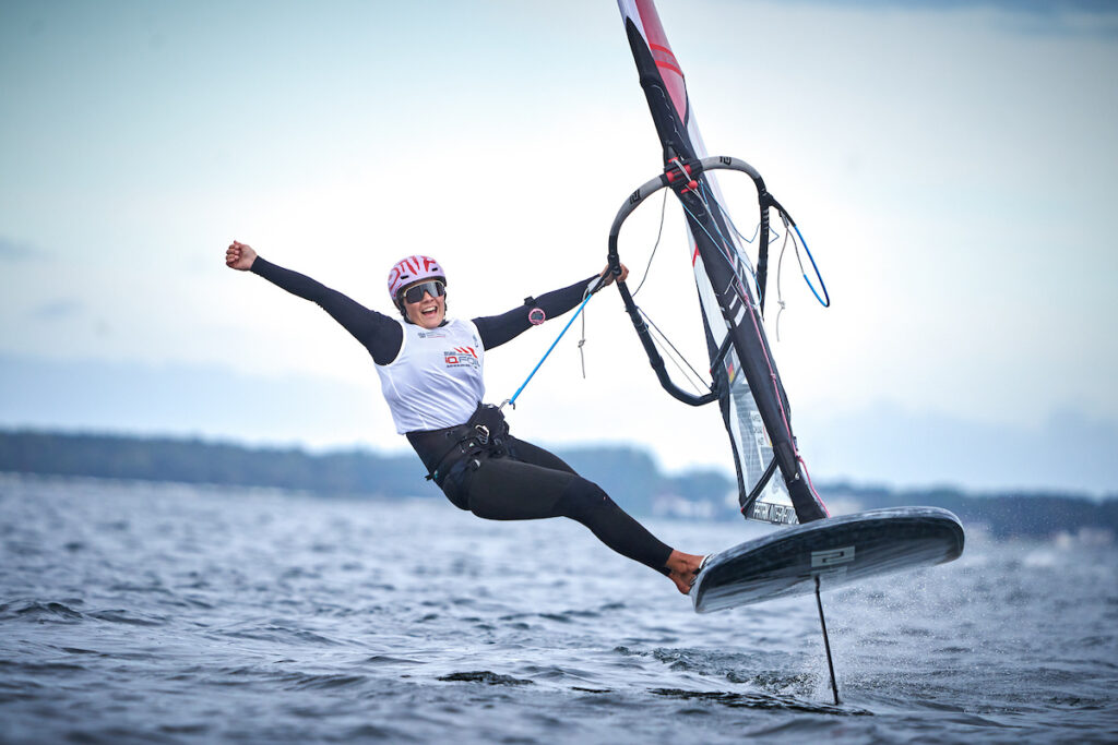 Eine Frau auf einem foilenden Surfboard streckt freudig den Arm in die Höhe.