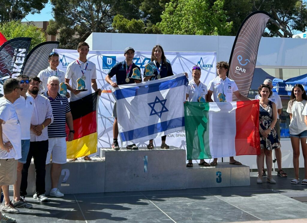 Auf dem Siegerpodest stehen drei Segelteams mit ihren Nationalflaggen. In der Mitte die israelische Flagge, links daneben die deutsche und rechts die italienische Flagge.