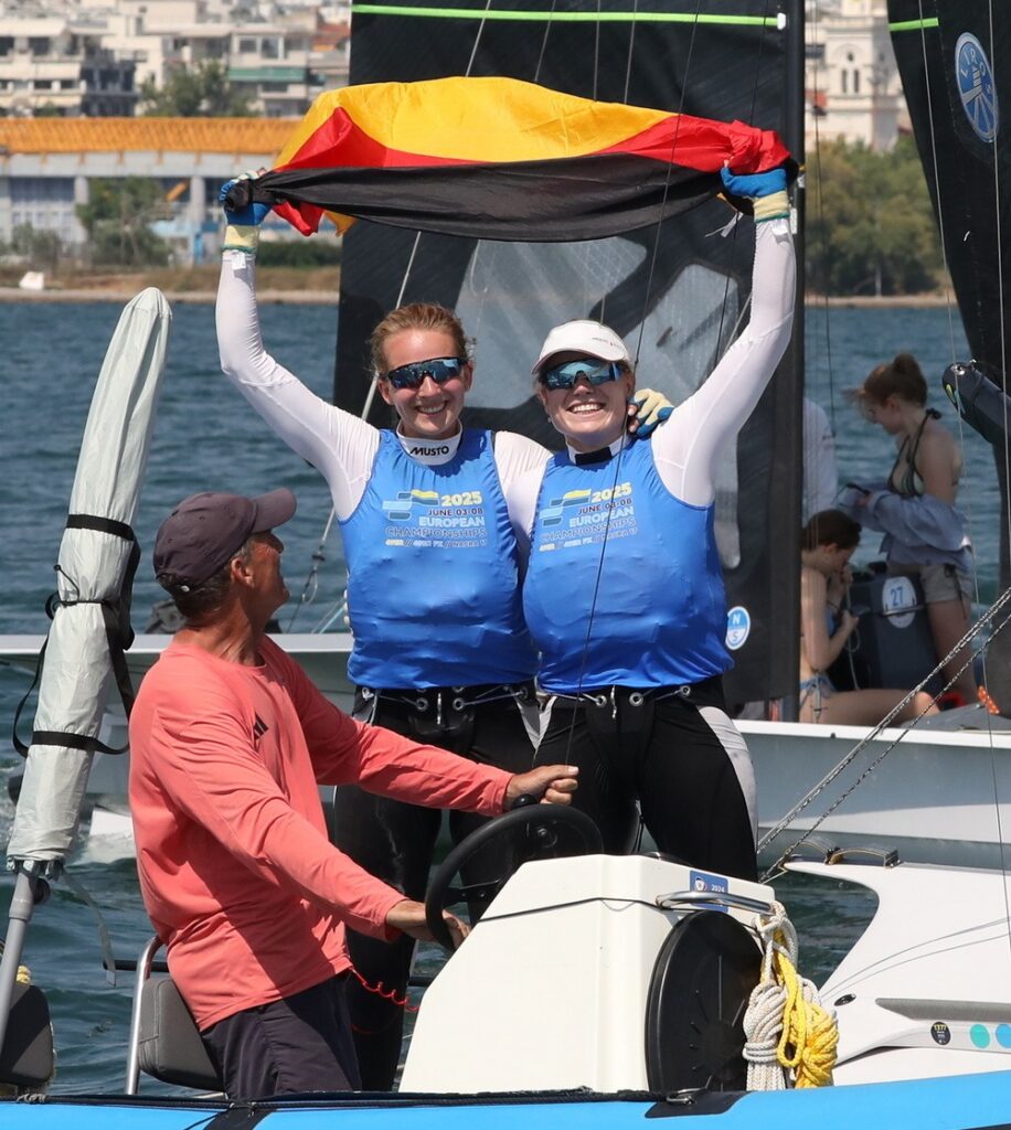 Marla Bergmann und Hanna Wille stehen im bauen Bib auf ihrem Boot und halten jubelnd eine Deutschlandflagge in die Luft. Vor ihnen sitzt ihr Coach Maurice Paardenkooper auf dem Motorboot