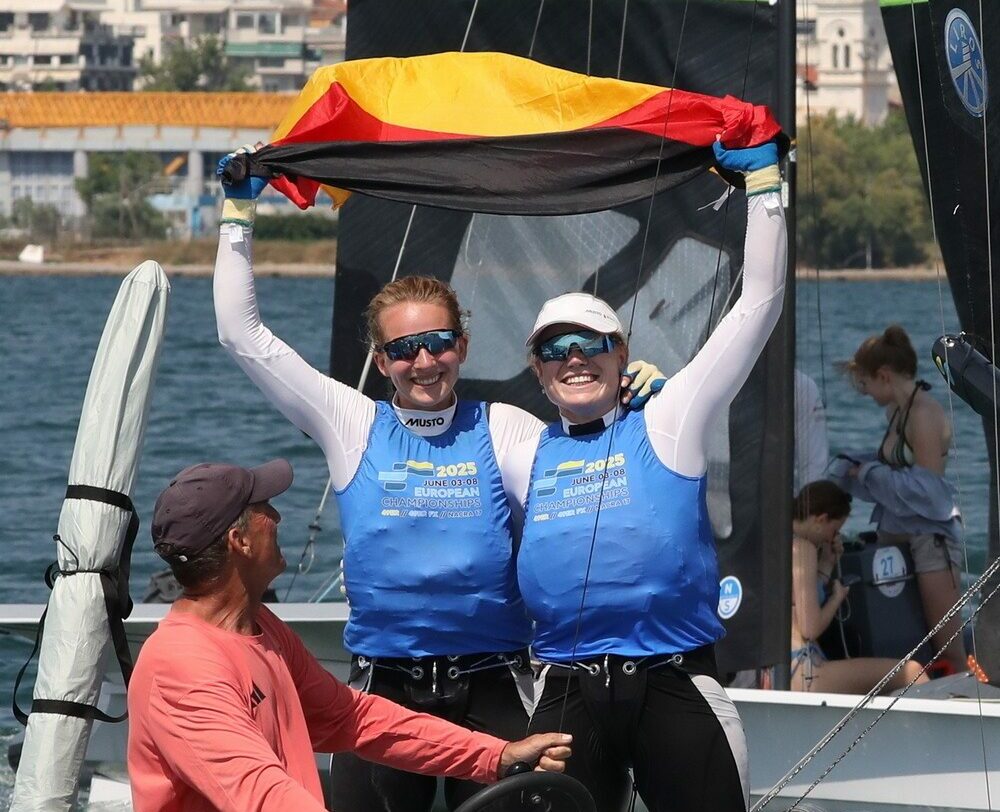 Marla Bergmann und Hanna Wille stehen im bauen Bib auf ihrem Boot und halten jubelnd eine Deutschlandflagge in die Luft. Vor ihnen sitzt ihr Coach Maurice Paardenkooper auf dem Motorboot